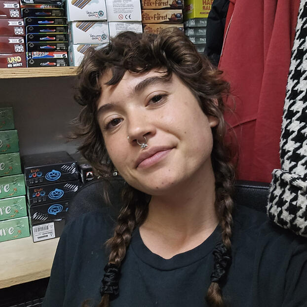Corvus, a person with light skin, braided curly brown hair, and a calm smile. The image is of their head, slightly tilted, looking at the camera.